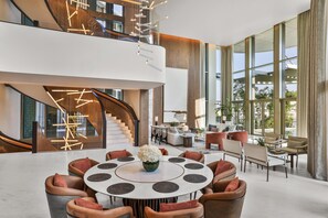 A spacious lobby with a central round table, seating arrangements, a staircase, and large windows.