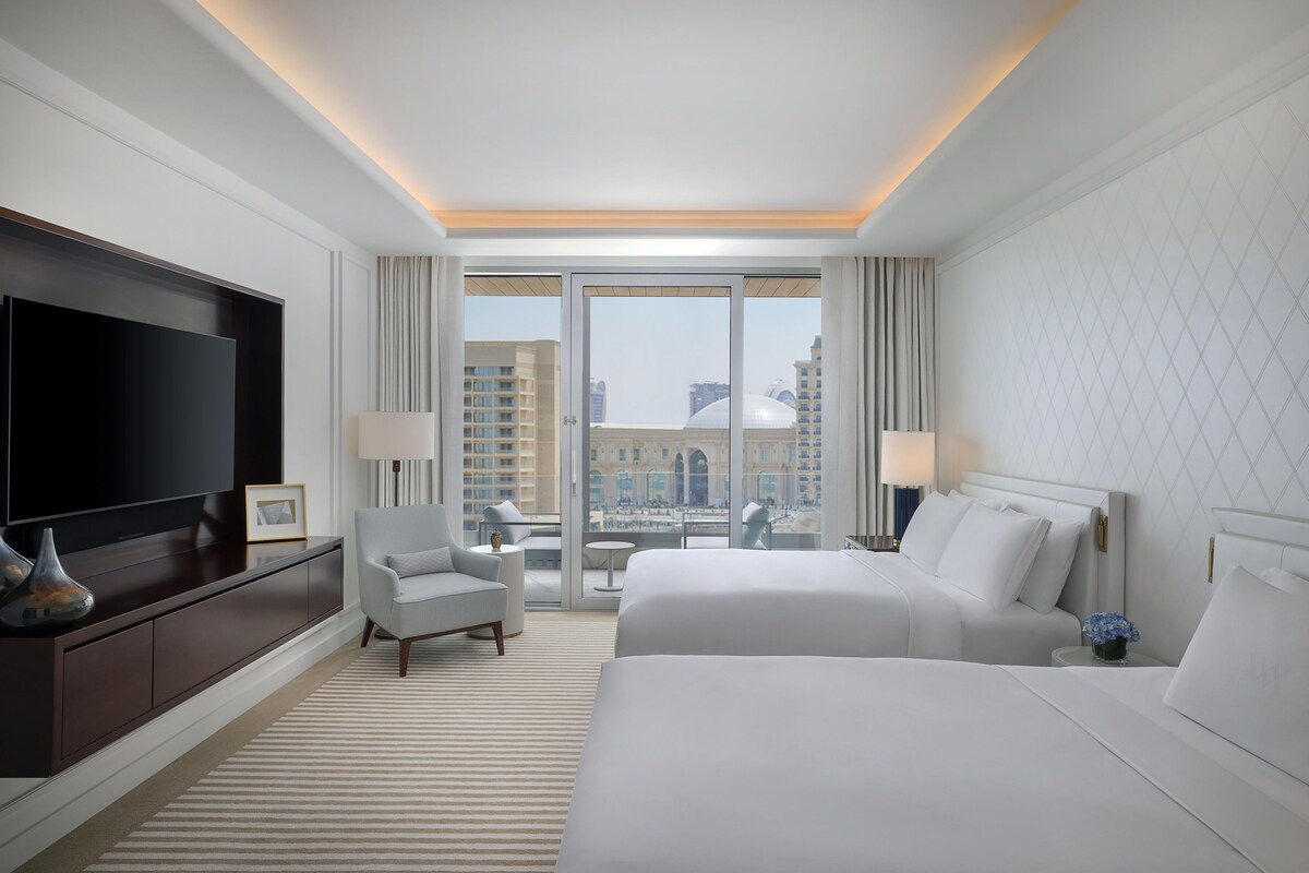 A hotel room with a large TV, two beds, a sofa, a small table, and a view of the city through large windows.