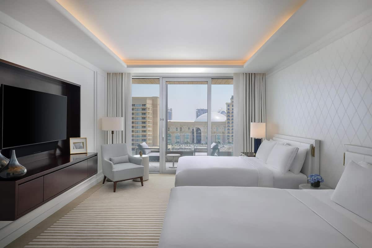 A hotel room with a large TV, two beds, a sofa, a small table, and a view of the city through large windows.