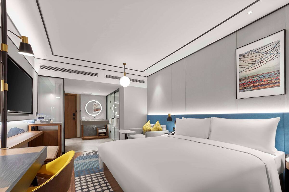A modern hotel room with a large bed, a desk, a chair, a bathroom with a mirror, and a painting on the wall.