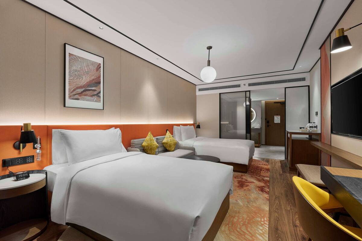 A hotel room with two beds, a desk, a chair, a TV, a clock, and a painting on the wall.