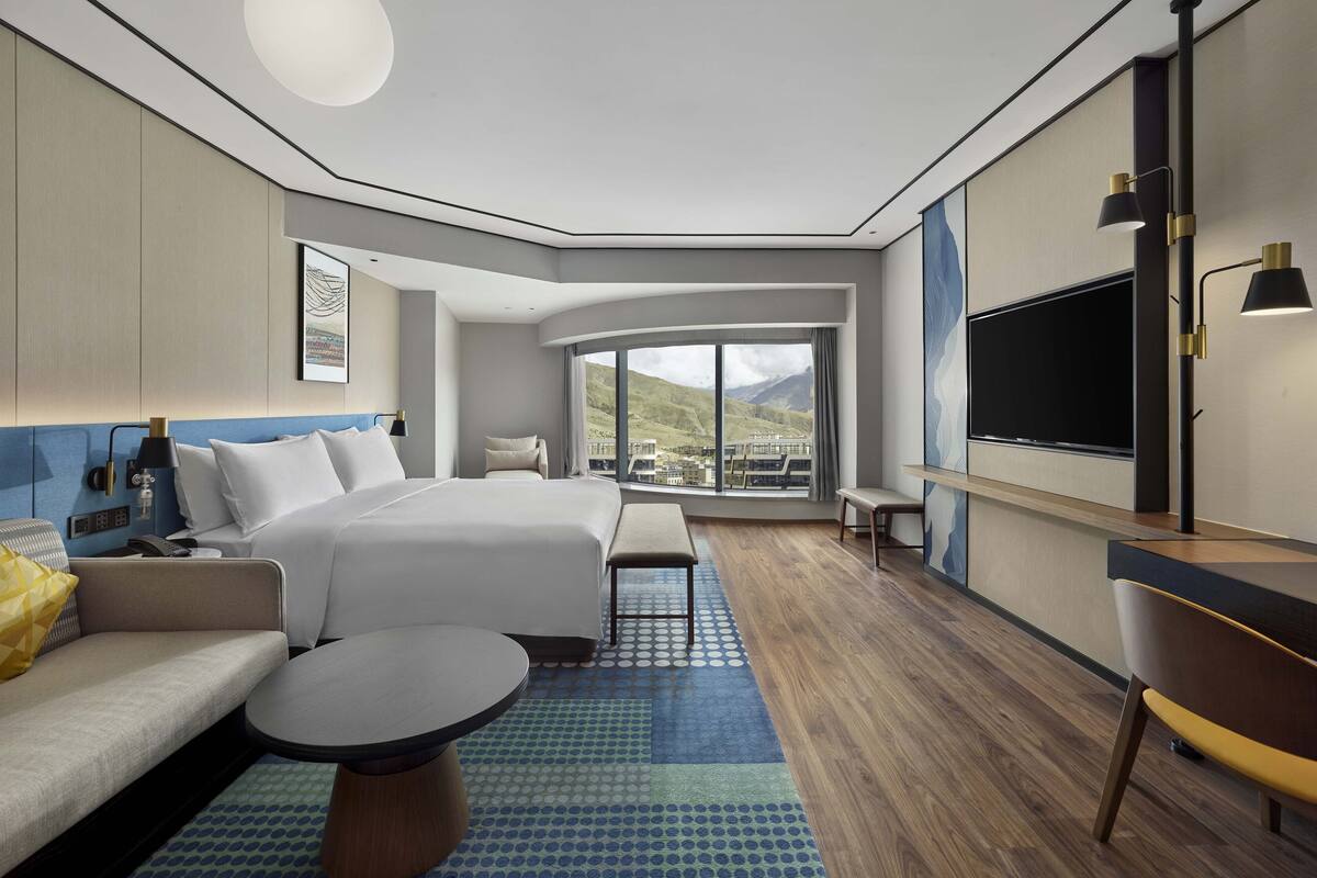 A modern hotel room with a large bed, a sofa, a dining table, and a TV.