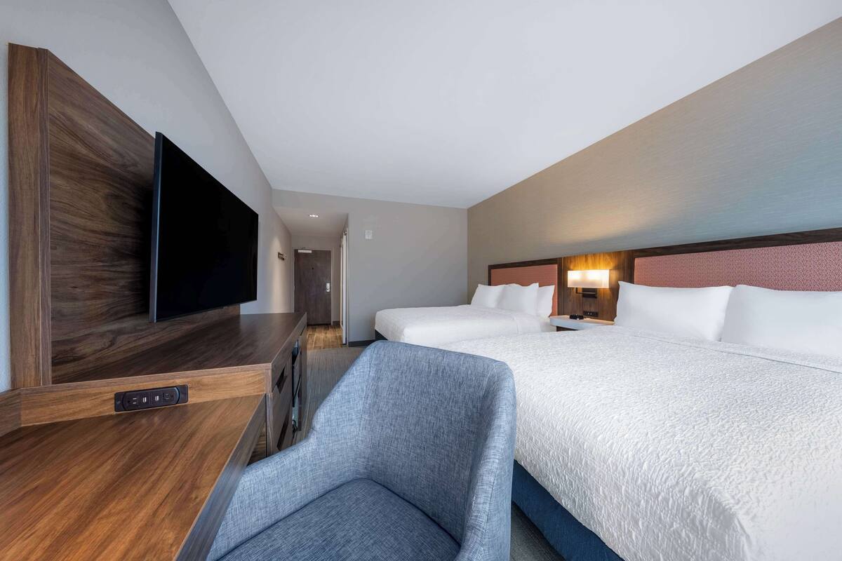 Habitación de hotel con una televisión de pantalla plana montada en un panel de madera, una butaca tapizada en gris, dos camas con ropa blanca y un cabecero con iluminación integrada.