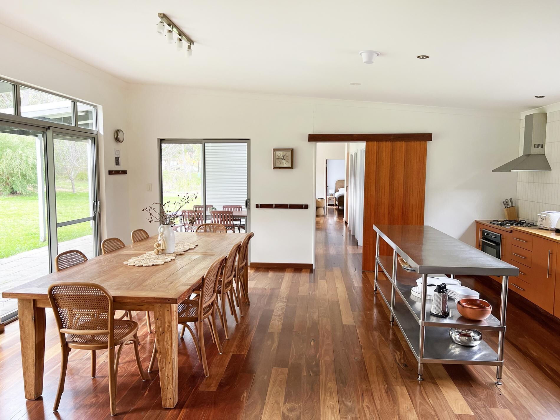 A modern kitchen with a long dining table, wooden flooring, and a view of the outdoors.