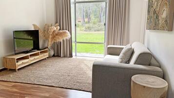 A modern living room with a grey sofa, a wooden TV stand, a large window with curtains, and a view of a grassy area.