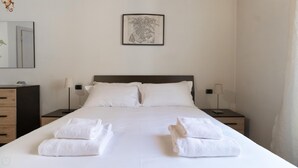 A neatly made bed with white linens and towels, two bedside tables with lamps, a framed map on the wall, and a wooden nightstand with a lamp. - Italianway Easy - Muratori 8 (Milan)