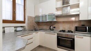 A modern kitchen with white cabinets, a stainless steel range hood, a microwave, an oven, and a window with blinds. - Italianway Easy - Muratori 8 (Milan)