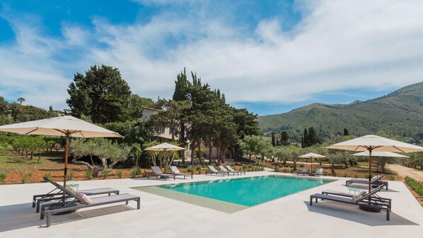 Zona de piscina con sillones de descanso y sombrillas, rodeada de árboles y una montaña al fondo.
