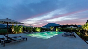 Área de piscina com espreguiçadeiras e guarda-sóis, cercada por vegetação e montanhas ao fundo.