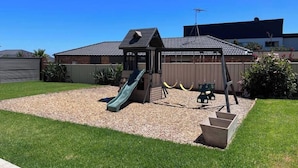 Een speelplaats met een glijbaan en schommels, omringd door een omheinde tuin met een grindpad en gras.