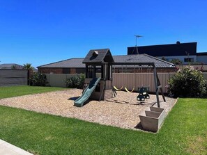 A playground with a slide and swings, surrounded by a fenced yard with a gravel area and grass.