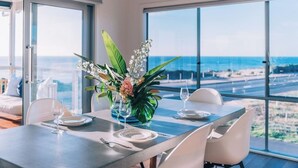 Un coin repas avec une table dressée pour deux personnes, de grandes fenêtres offrant une vue sur l’océan et une plante en pot ornée de fleurs blanches.