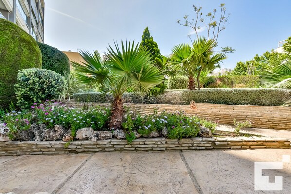 A landscaped garden with a variety of plants, including palm trees and flowering shrubs, bordered by a stone wall.