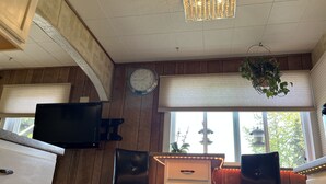 Une pièce aux murs recouverts de lambris en bois, avec une horloge blanche, un téléviseur, deux chaises, un petit meuble blanc et une fenêtre avec des stores.