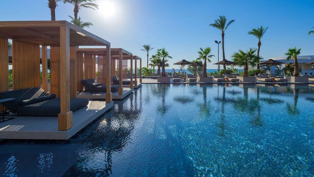 A poolside area with lounge chairs and umbrellas, palm trees, and a clear view of the ocean. - Ammades All Suites Beach Hotel & Spa - Adults Only (Rhodes)