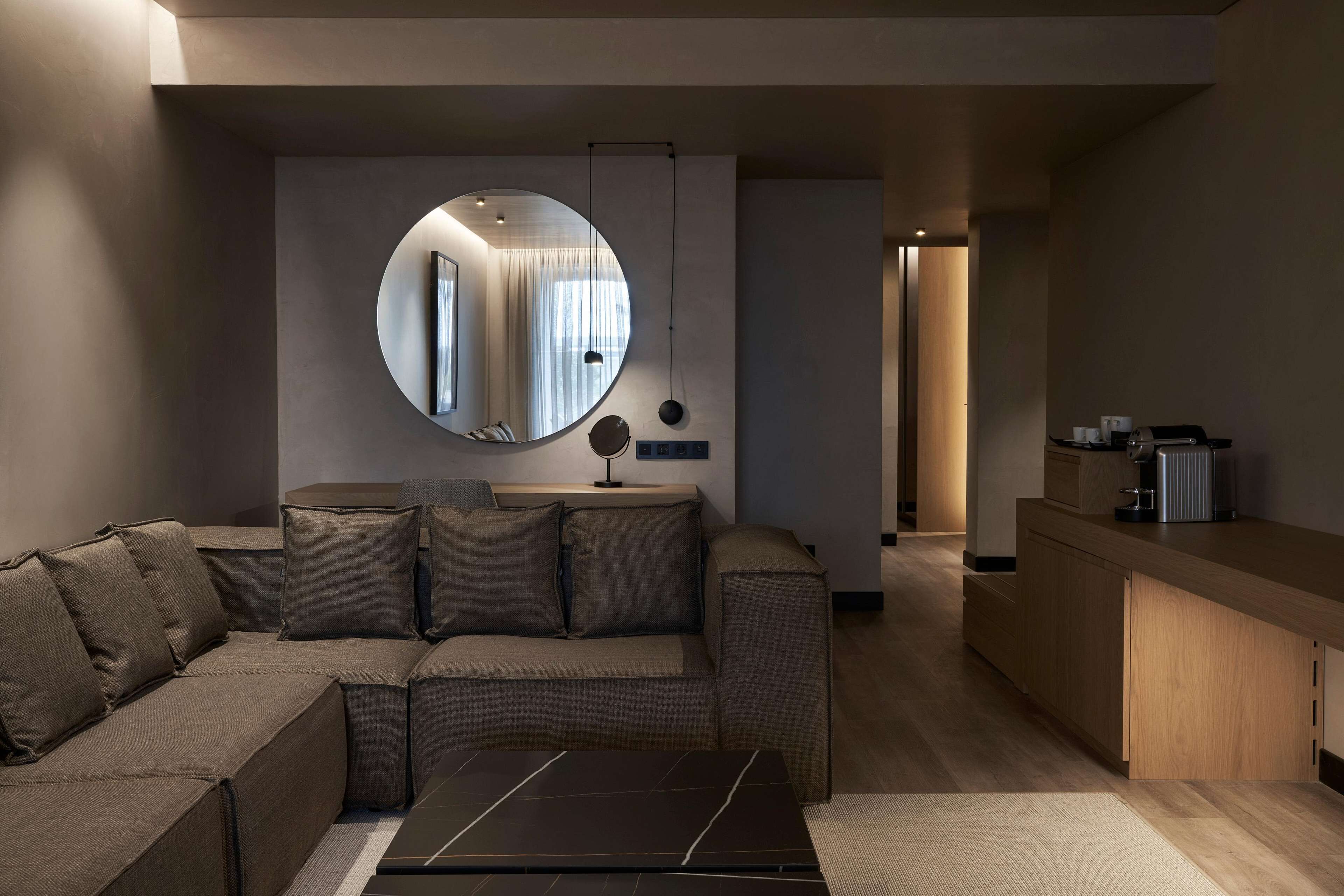 Una sala de estar moderna con un sofá modular, un espejo redondo, una mesa de centro y una zona de cocina con encimera y armarios.