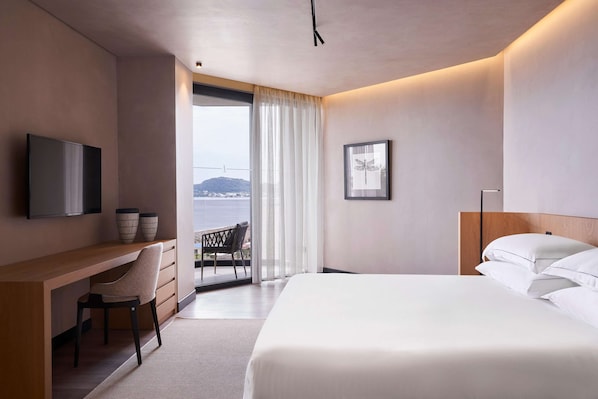 A modern hotel room with a bed, a desk, a chair, a TV, and a balcony with a view of the sea. - Ammades All Suites Beach Hotel & Spa - Adults Only (Rhodes)