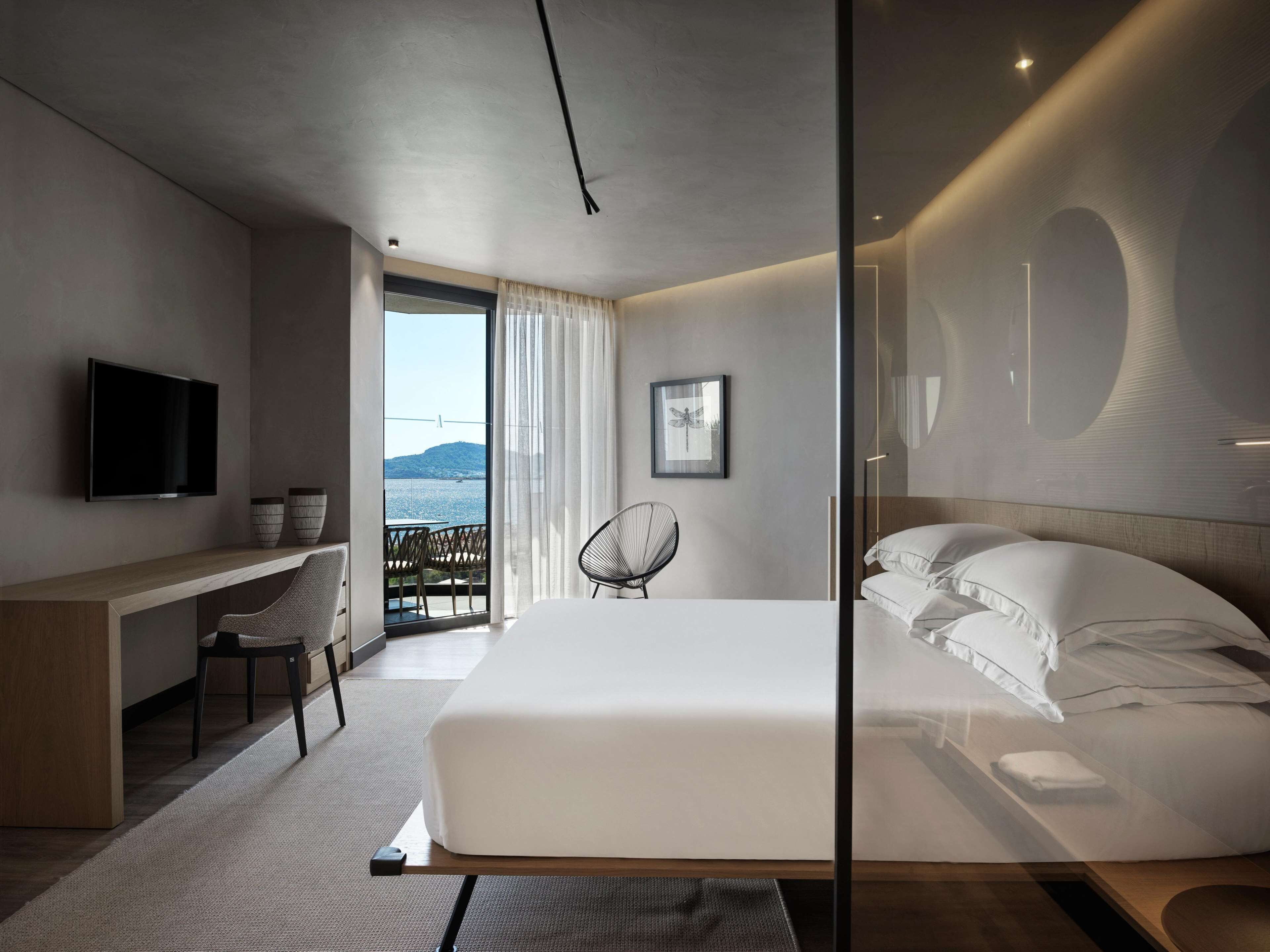 Habitación de hotel moderna con una cama grande, un escritorio, una silla, un televisor y un balcón con vista al mar.