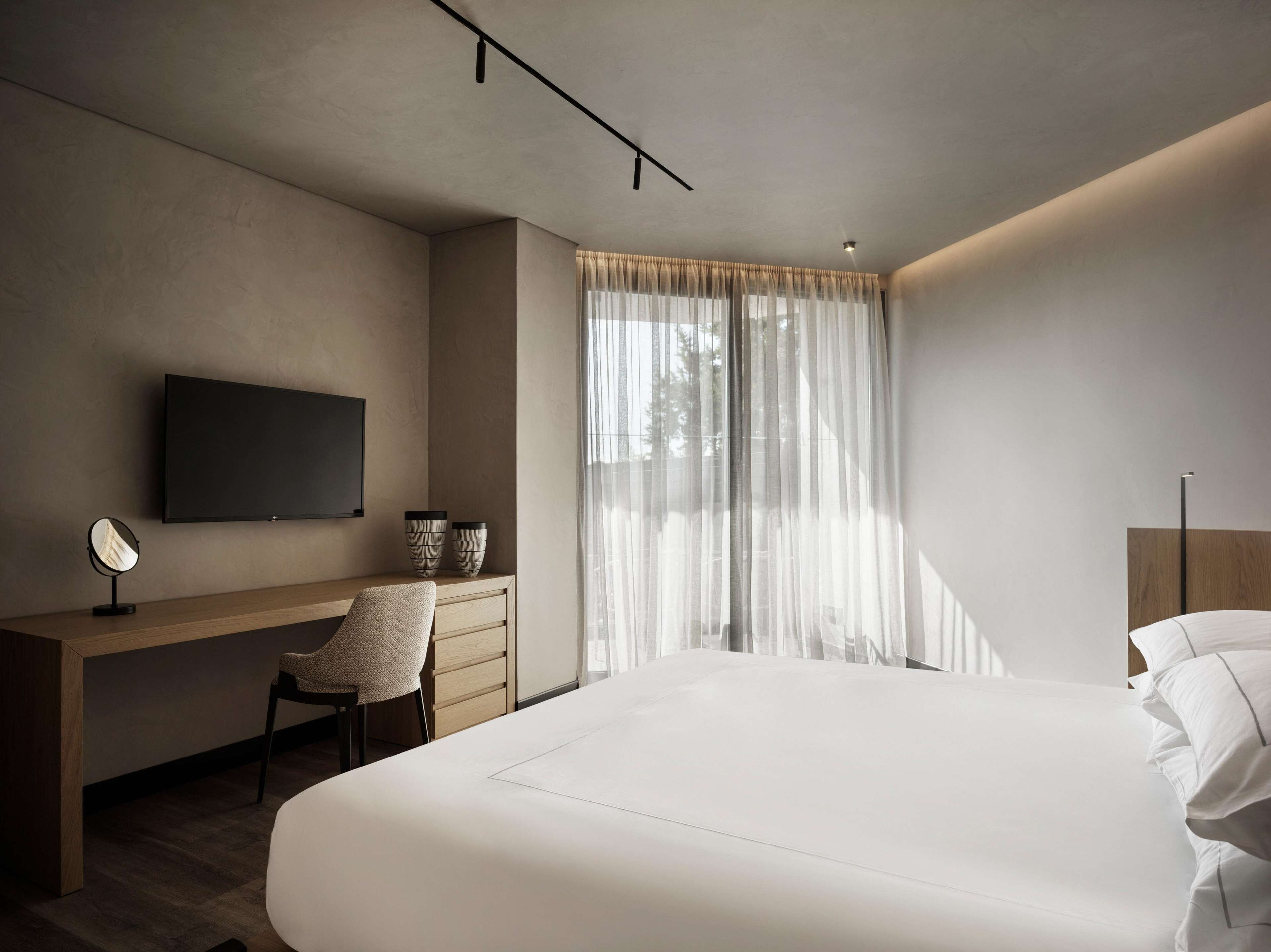 Un dormitorio moderno con una cama grande, un escritorio con silla, un televisor y una ventana con cortinas translúcidas.