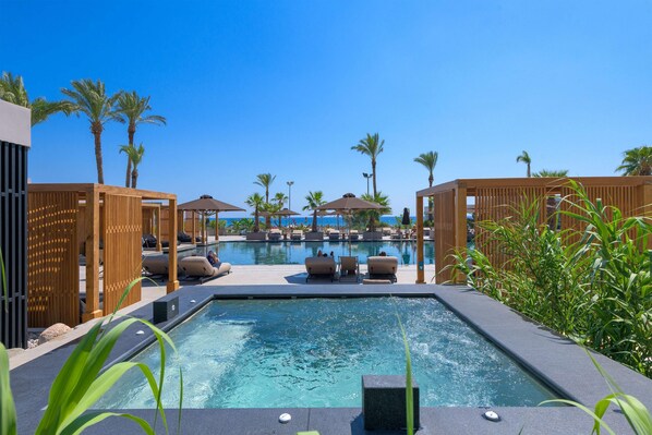 A pool area with lounge chairs and umbrellas, surrounded by palm trees and wooden structures. - Ammades All Suites Beach Hotel & Spa - Adults Only (Rhodes)