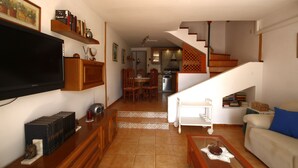 Una sala de estar con televisión de pantalla plana, estanterías de madera, zona de comedor con sillas, una escalera y una cocina al fondo.