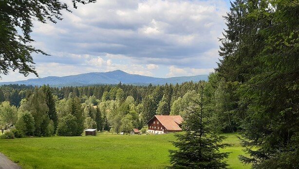 Un paysage rural avec une grange rouge, une route dégagée et une zone boisée, avec des montagnes en arrière-plan.