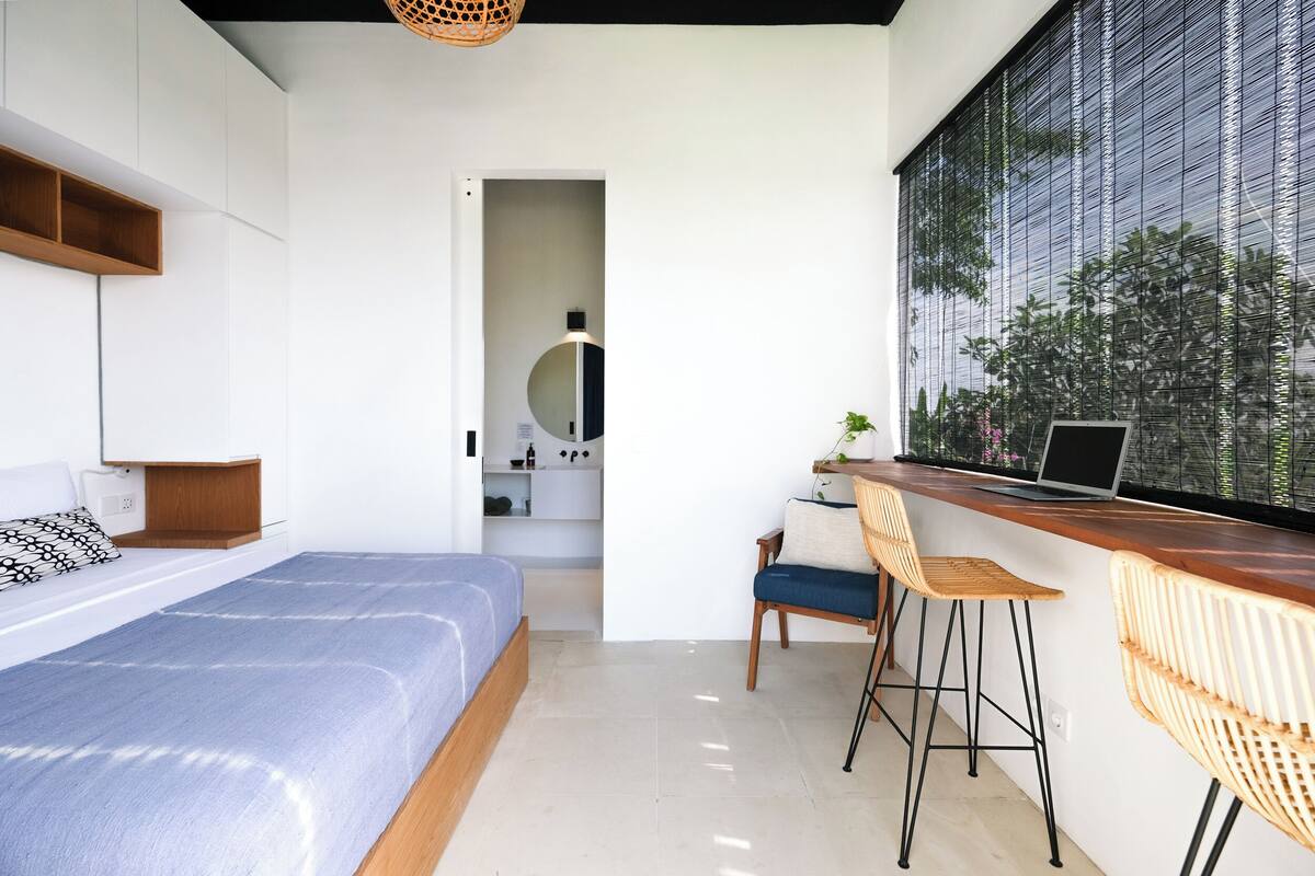 Un dormitorio moderno con cama, escritorio con computadora portátil y un amplio ventanal con persianas.