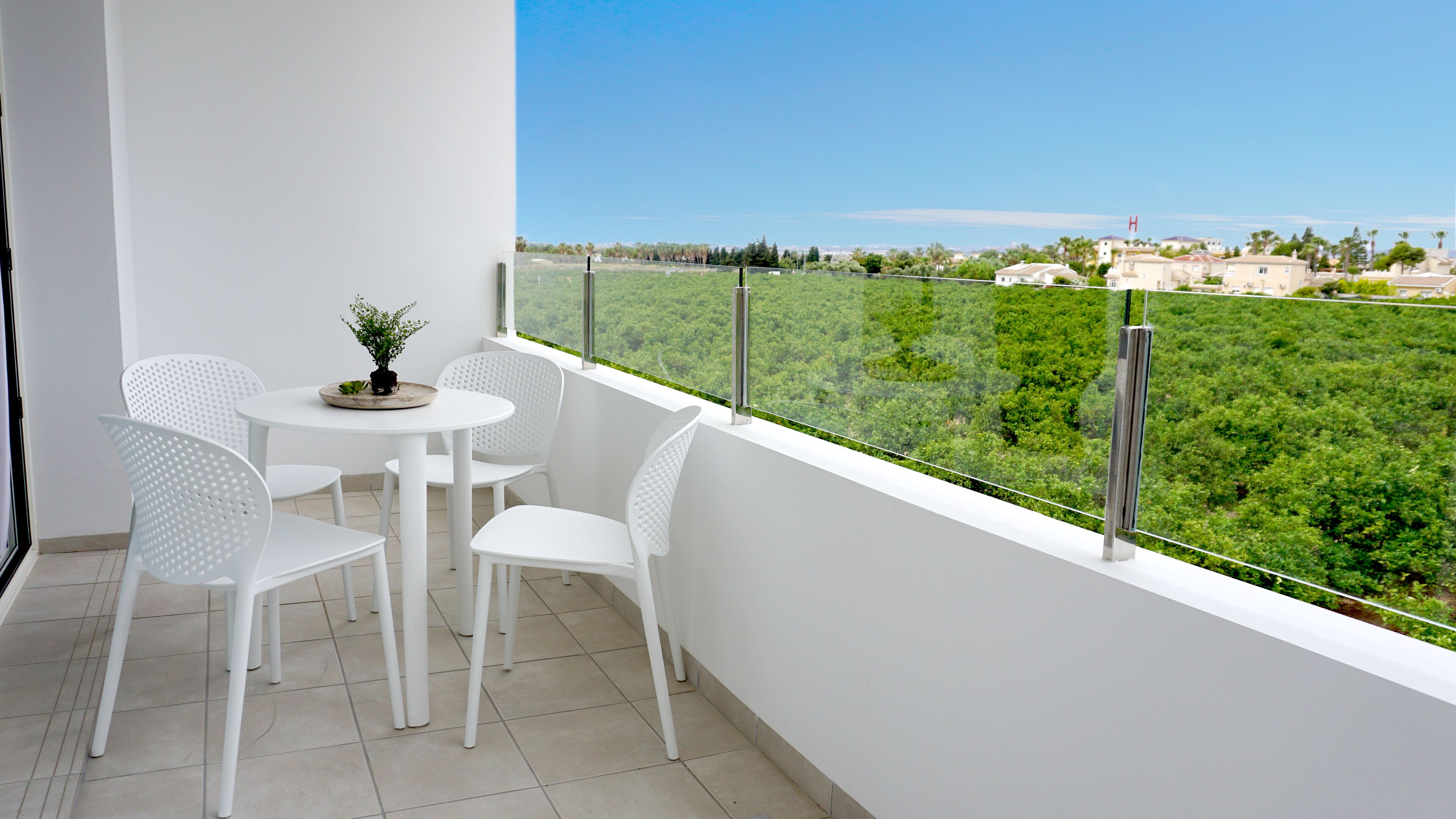 Ein Balkon mit weißen Stühlen und einem kleinen Tisch, mit Blick auf eine grüne Landschaft und Gebäude in der Ferne.