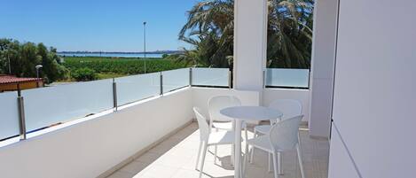 A balcony with white chairs and a table, overlooking a coastal landscape with palm trees and a clear sky.