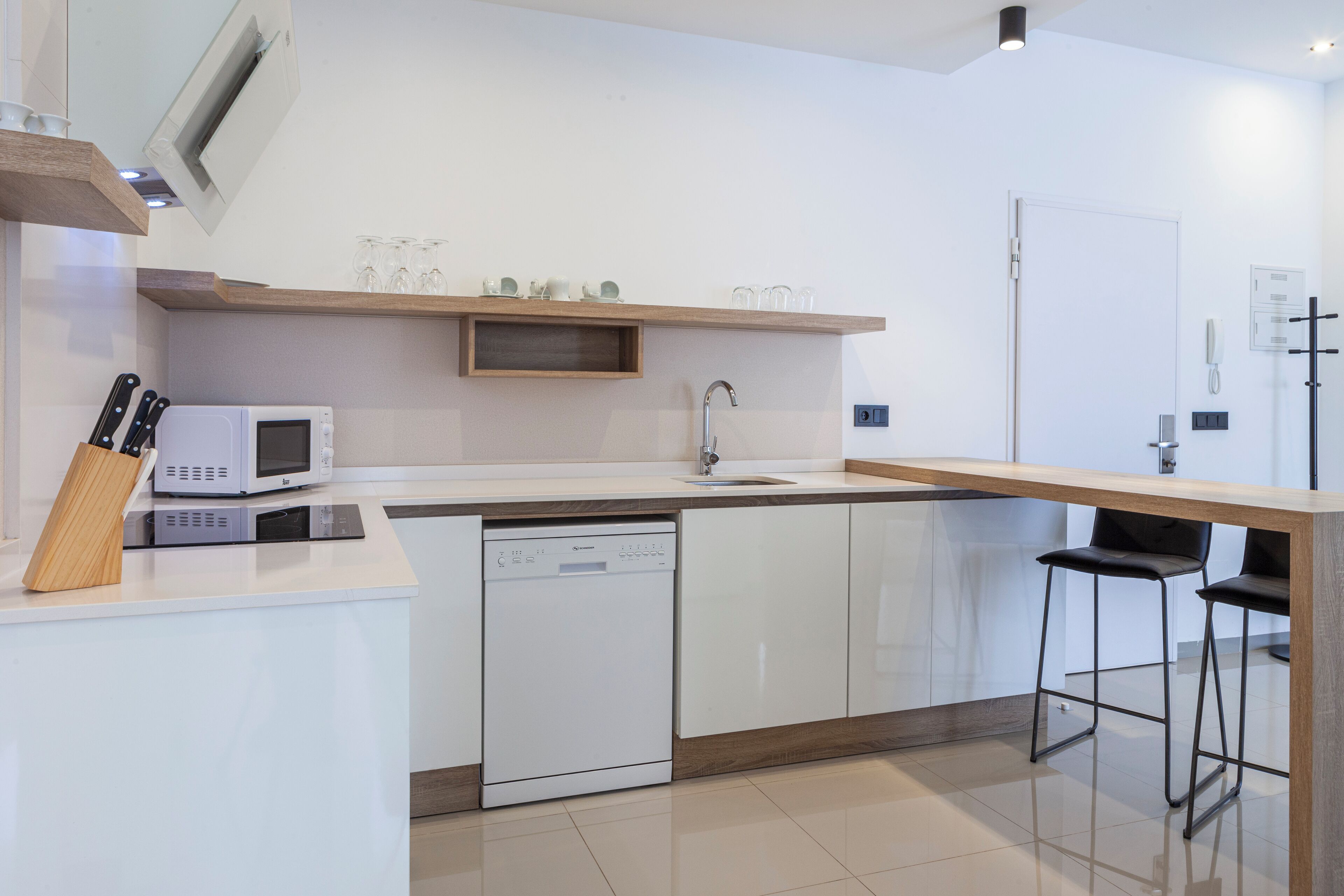 Una cocina moderna con armarios blancos, microondas integrado, lavavajillas y encimera de madera. Hay dos taburetes negros y un estante de madera sobre el fregadero.