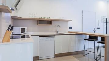 Una cocina moderna con armarios blancos, microondas integrado, lavavajillas y encimera de madera. Hay dos taburetes negros y un estante de madera sobre el fregadero.