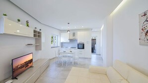 Una cucina moderna con mobili bianchi, un'area pranzo con tavolo e sedie bianche e un soggiorno con divano color beige.