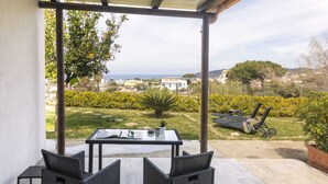 Un portico coperto con tavolo e sedie, affacciato su un giardino che si apre su una vista del mare e case in lontananza.