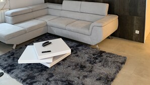 Un salon moderne avec un canapé d’angle gris clair, un tapis gris foncé à poils longs et une table basse blanche avec des télécommandes.