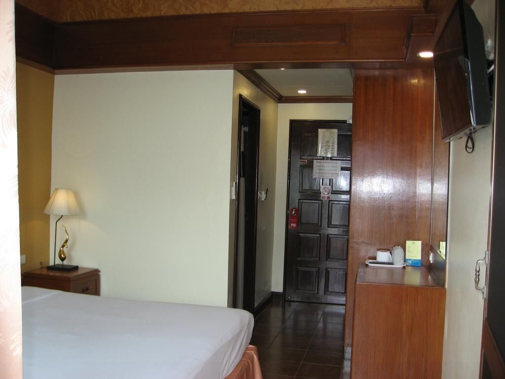 A hotel room with a bed, a nightstand with a lamp, a wooden wardrobe, and a kitchenette area with a refrigerator and a microwave.
