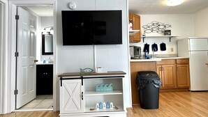 A modern kitchen with a flat-screen TV mounted above a white cabinet, wooden cabinets, a black trash bin, and a wooden floor.