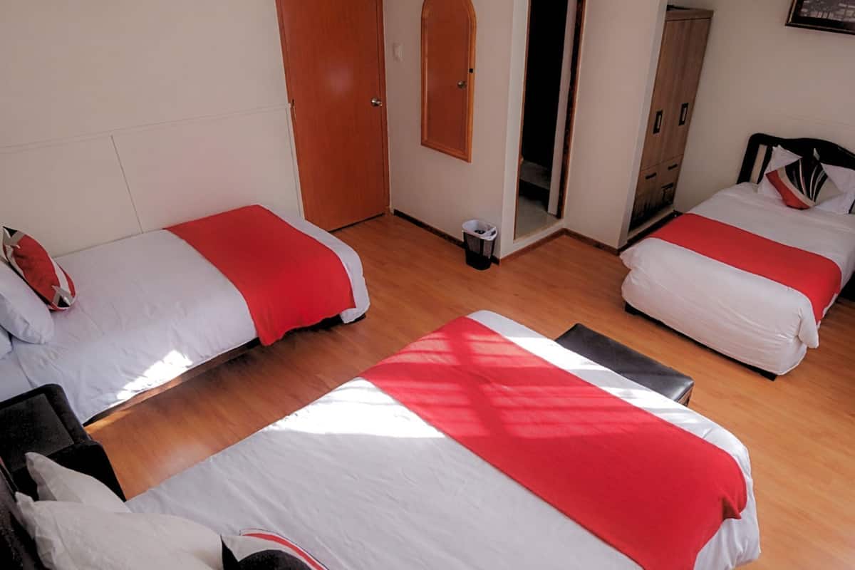 Habitación de hotel con dos camas, cada una con cubrecama rojo y blanco, suelo de madera, armario de madera y una mesita con lámpara.