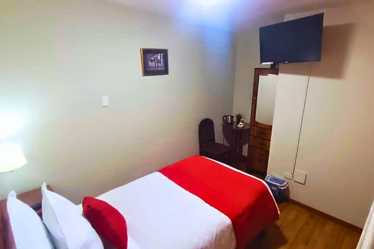 Una habitación pequeña y sencilla con una cama, un escritorio, una silla, un televisor y un espejo.