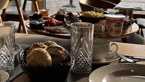 Una mesa de comedor con diversos alimentos, como pan, frutas y yogur, ubicada en una habitación con ventanas amplias que dan al exterior.