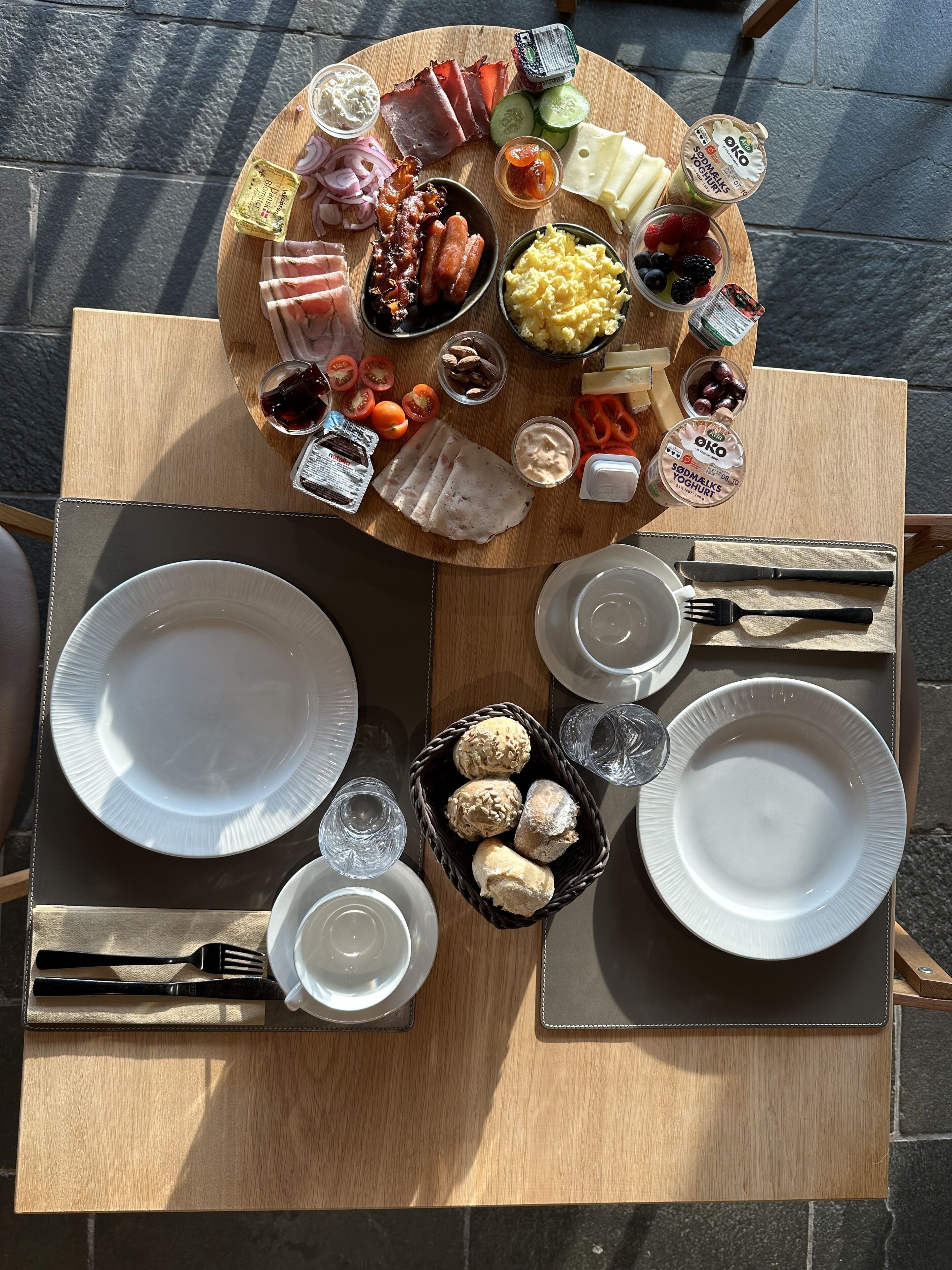 A wooden table with a variety of food items including meats, cheeses, fruits, and yogurt, along with plates and cutlery.