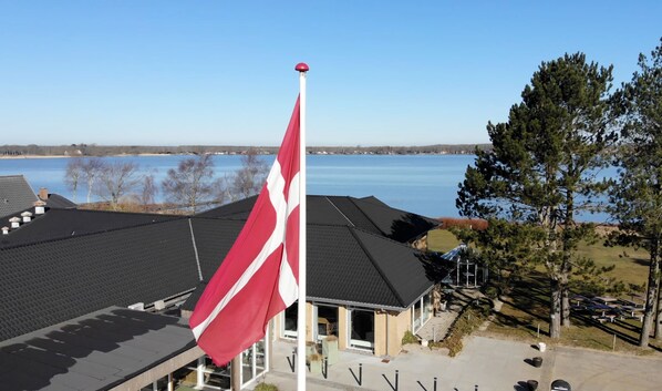 Un asta con una bandera, un edificio de techo oscuro y vistas a un lago y árboles.