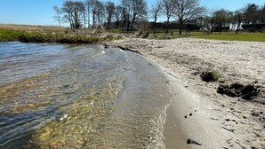 Una playa con aguas claras y poco profundas, orilla arenosa y árboles al fondo.