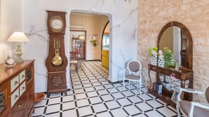 Um hall com piso de mármore, um relógio grande, um candelabro e uma parede de pedra.