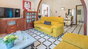 Uma sala de estar com um sofá amarelo, uma televisão, um armário de madeira e um chão ladrilhado.
