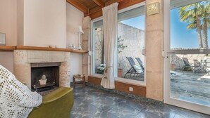 Um quarto aconchegante com lareira, uma poltrona verde e vista para um pátio externo com cadeiras e uma palmeira.