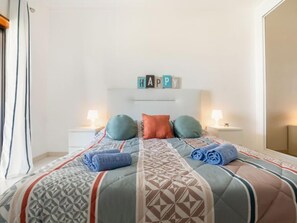 A neatly made bed with a patterned bedspread, pillows, and rolled towels. There are two bedside tables with lamps. Above the bed, there are blocks spelling "HAPPY".