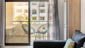 Een moderne woonkamer met een zwarte bank, een klein rond tafeltje met een hoed en fruit, en uitzicht op een balkon met stoelen en een gebouw op de achtergrond.