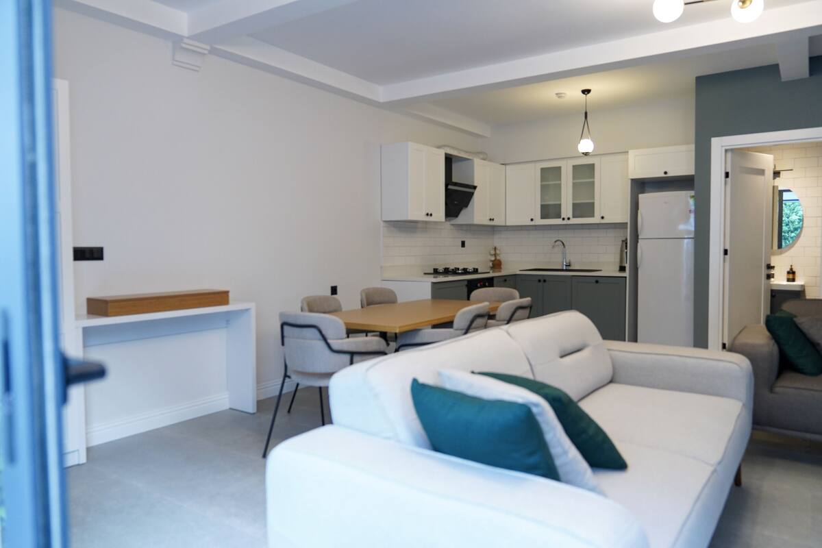 A modern kitchen with white cabinets, a dining area with a wooden table and chairs, and a white sofa with green cushions.