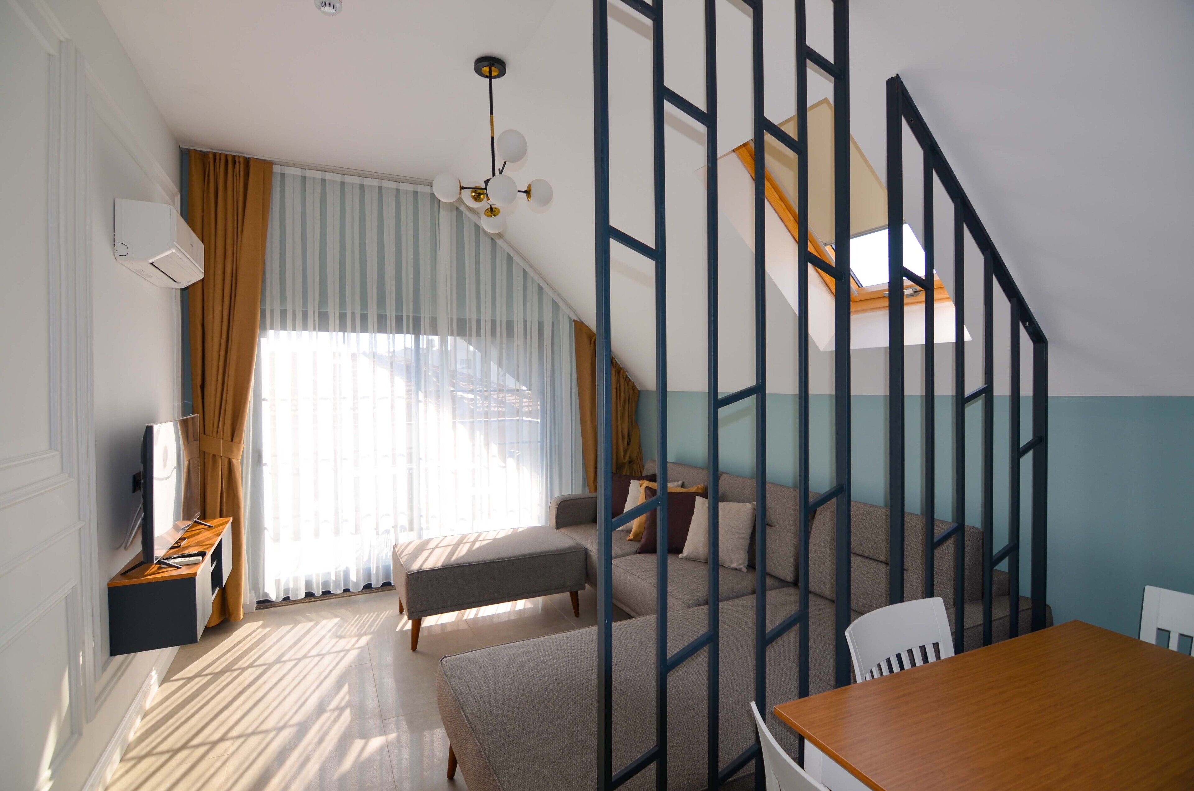 A modern living room with a sofa, ottoman, and a dining table. There is a staircase with a railing, and a window with curtains.