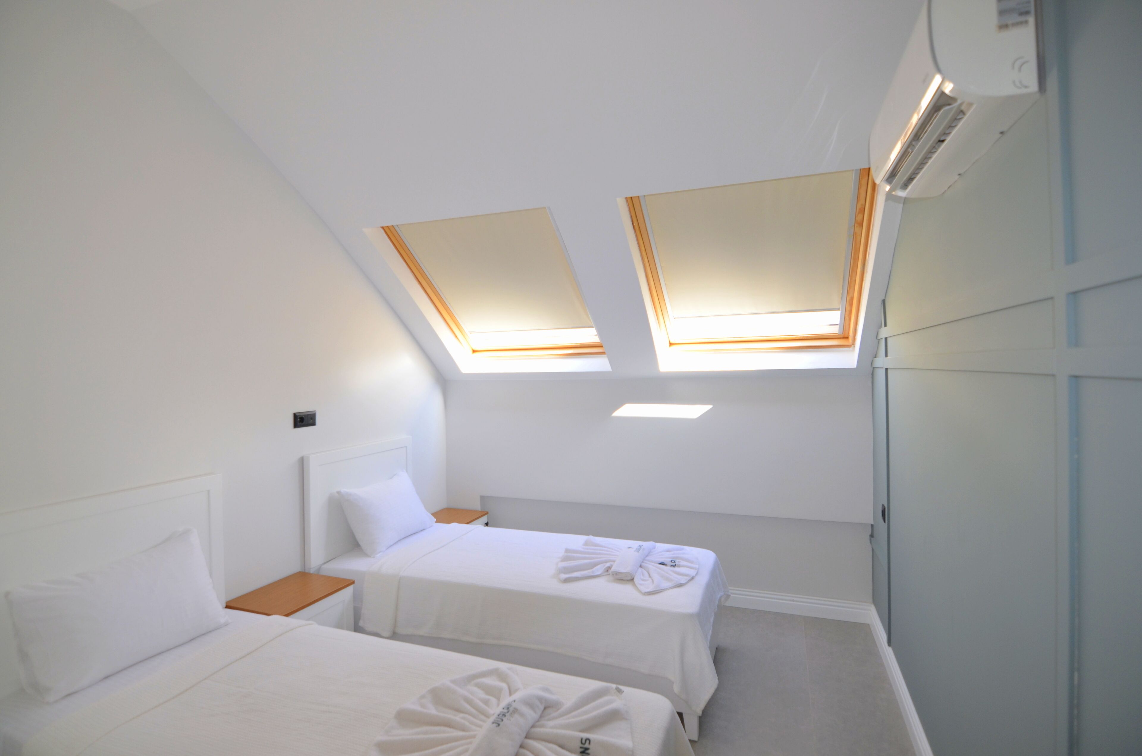A hotel room with two beds, skylights, an air conditioning unit, and a small wooden nightstand.